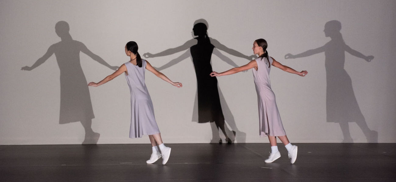 Two women in minimalist gray dresses and white sneakers dance onstage. Behind them three shadows echo their movement, of arms outstretches as in mid-stride.