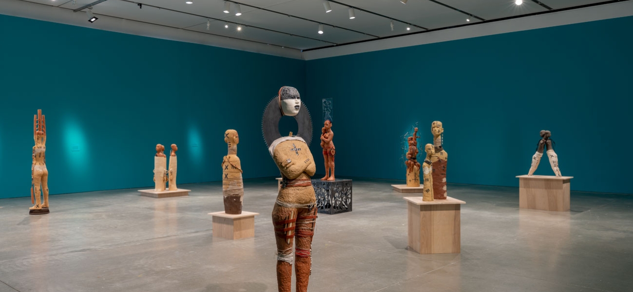 Clay figurative sculptures arranged in an open gallery, some standing on the floor and some on wooden plinths