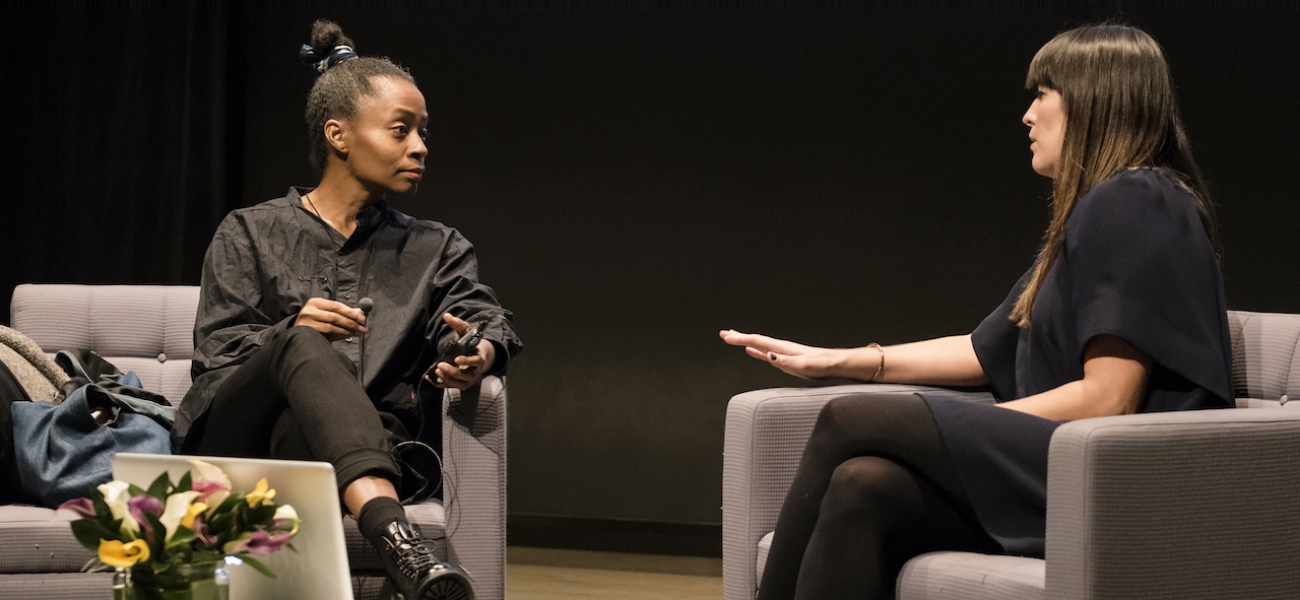 The artist Kara Walker and curator Eva Respini sit and talk onstage in gray chairs against a black background