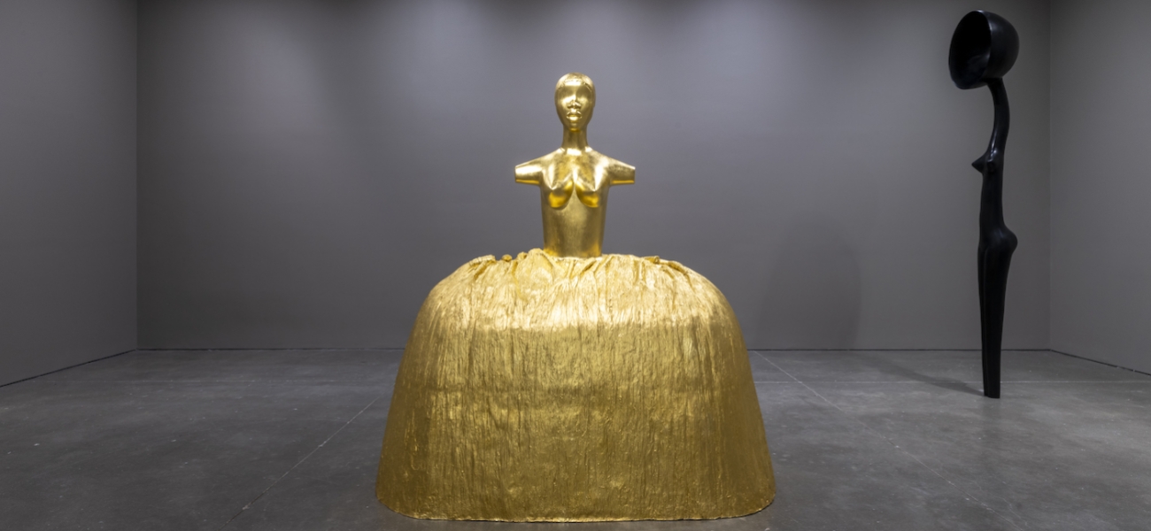 Two sculptures in a room with gray walls: a gold female figure with a voluminous skirt and an elongated, stylized female bronze with a satellite dish in place of a head. 