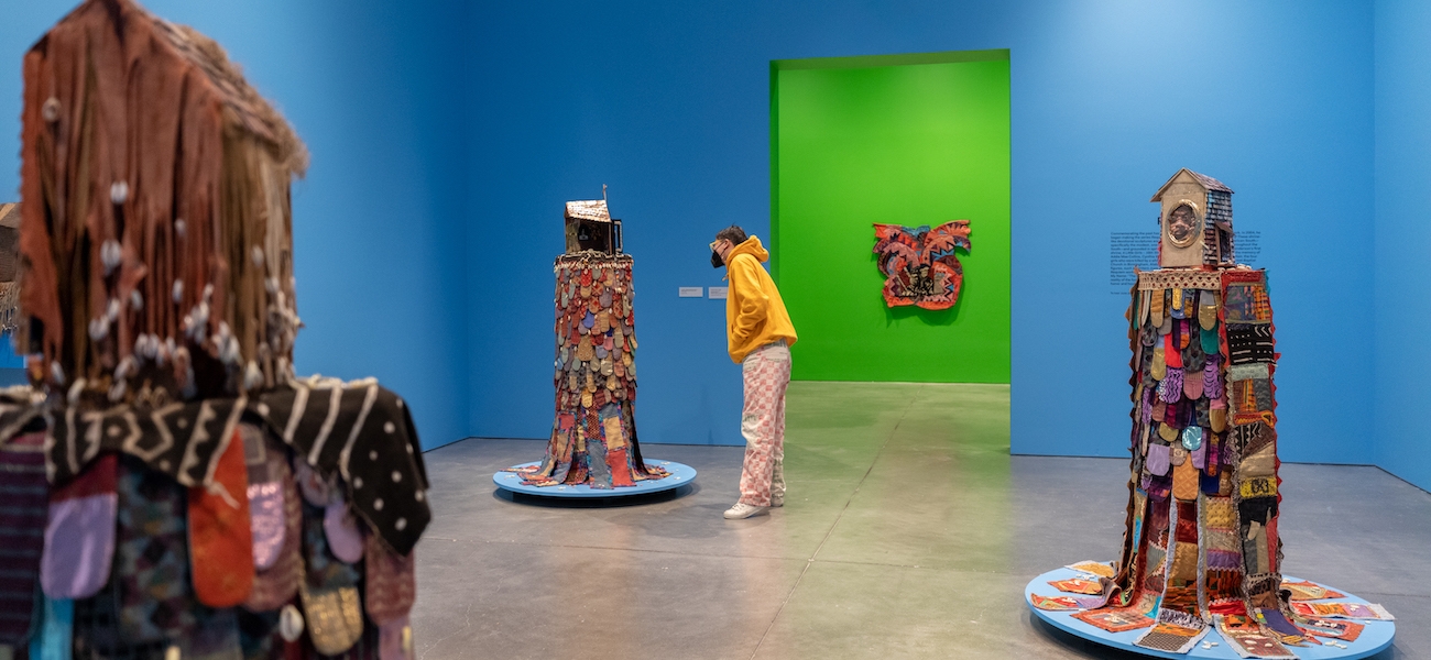 A person in a bright yellow hoodie looks at one of three standing mixed-media works in a gallery with bright blue walls. Tn the background is a gallery painted green with a colorful hanging work in view.