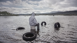 A person in a light windbreaker sits on a bucket in silvery water among abandoned tires.