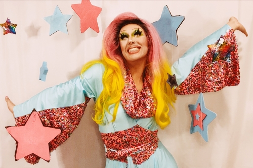 Photo of a drag queen in a pink and blue sparkly outfit posed against a starry garland