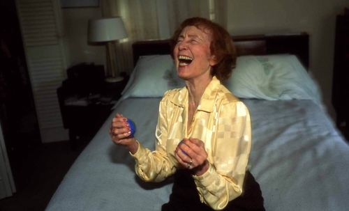 A color photograph of an older light-skinned woman wearing a yellow blouse and black pants and laughing widely while seated on the edge of a bed.