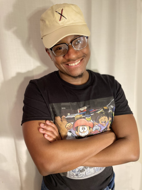 A man wearing a tan cap and a cartoon t-shirt smiling with his arms crossed