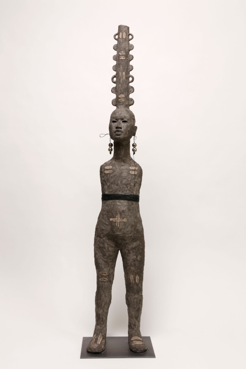 An armless grey clay figure with a cylindrical long opening on its head with multiple handles and bumps along it