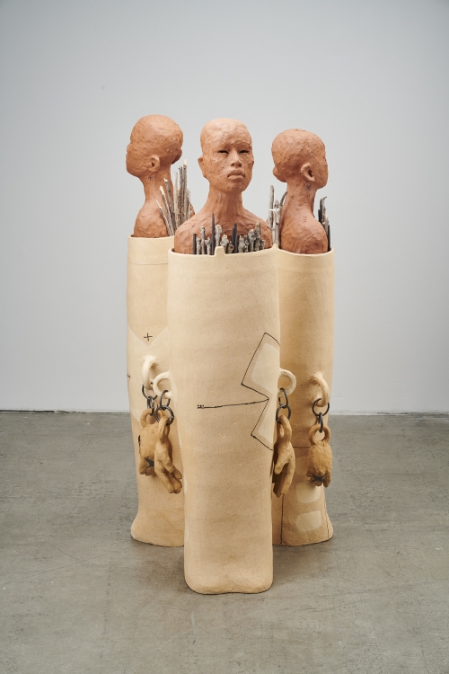 Three bald clay figures are wrapped tightly in a covering with sticks and rods protruding out