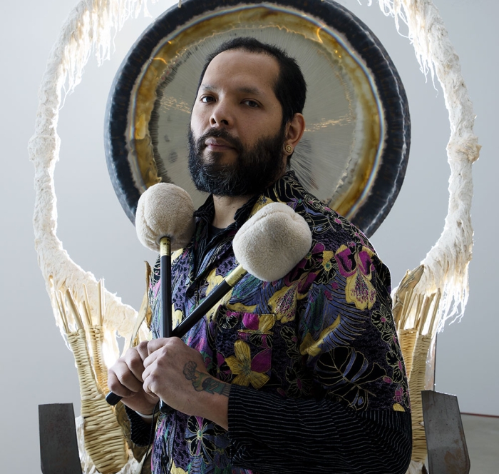 Artist Guadelupe Maravilla standing front of a gong sculpture holding two felted mallets 