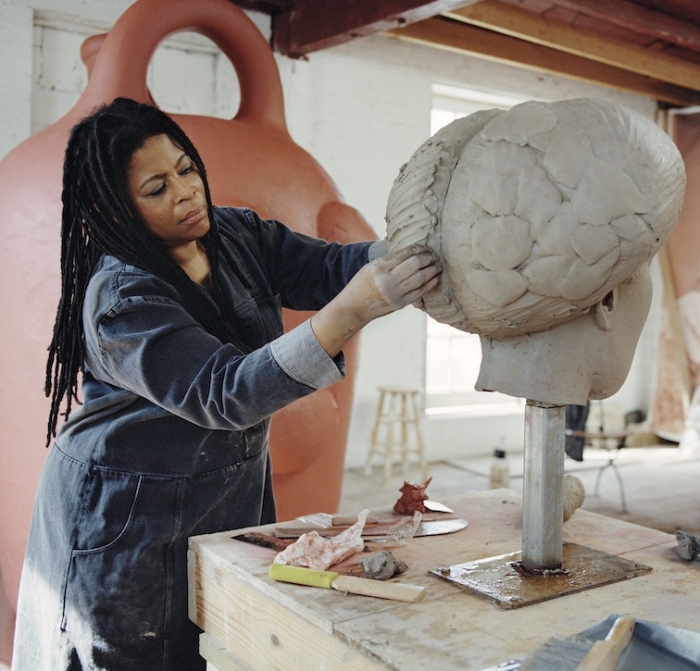 Simone Leigh, a medium-dark-skinned woman with dreadlocks in a denim work outfit, works on a large clay sculpture. 