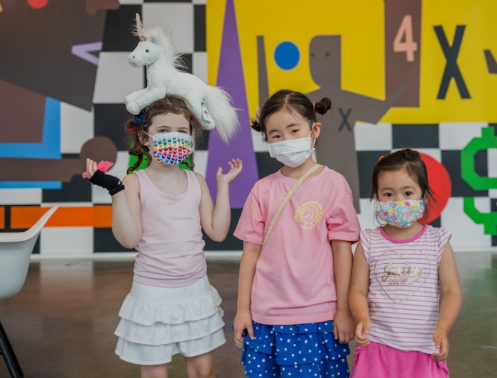 Three children wearing colorful masks and standing in front of the art wall. 