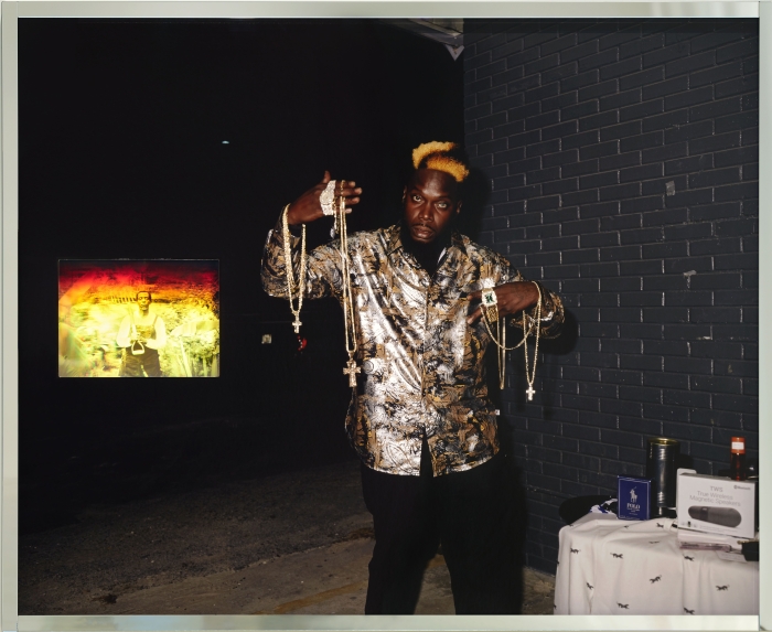 A Black man in a gold and black outfit poses with golden necklaces he is selling, draped from his arms. A hologram of an older Black man in overalls in a farm setting is embedded into the work.