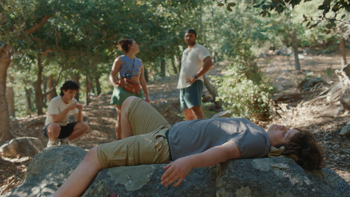 A boy lying down on some rocks outside with eyes closed with a few people standing in the distance