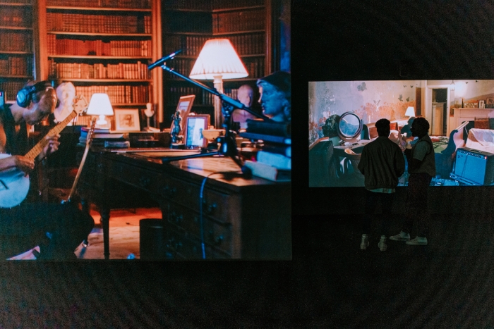 A large video screen of a musician with headphones and a guitar, and two visitors in the background watching a separate screen.