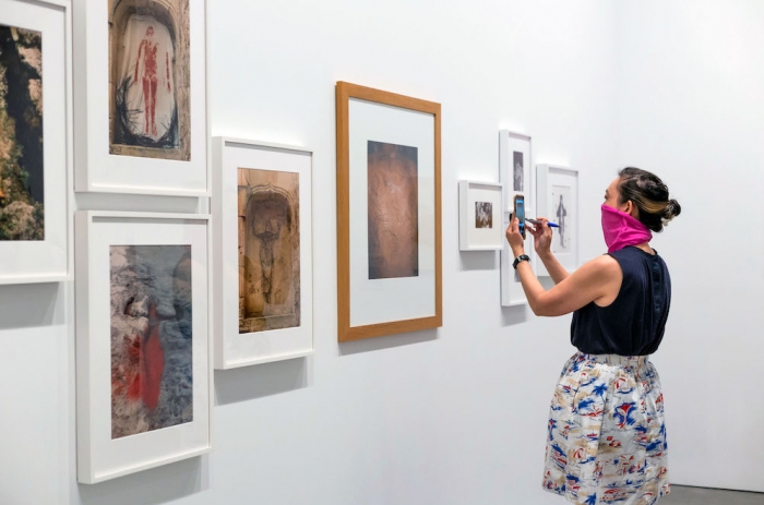 A young woman in a face covering takes a cell phone photo of a wall of artworks by Ana Mendiata, Mona Hatoum, and others in the exhibition "Beyond Infinity: Contemporary Art after Kusama."