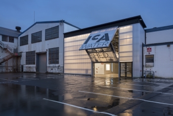A building with a translucent front wall and garage-door like opening reading "ICA Watershed" seems to glow from within in an industrial area at dusk.