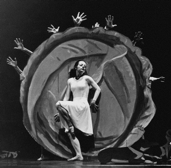 Black and white photo of a female dancer with light skin leaning expressively against a large cabbage painting, surrounded by several pairs of hands reaching outwards. 