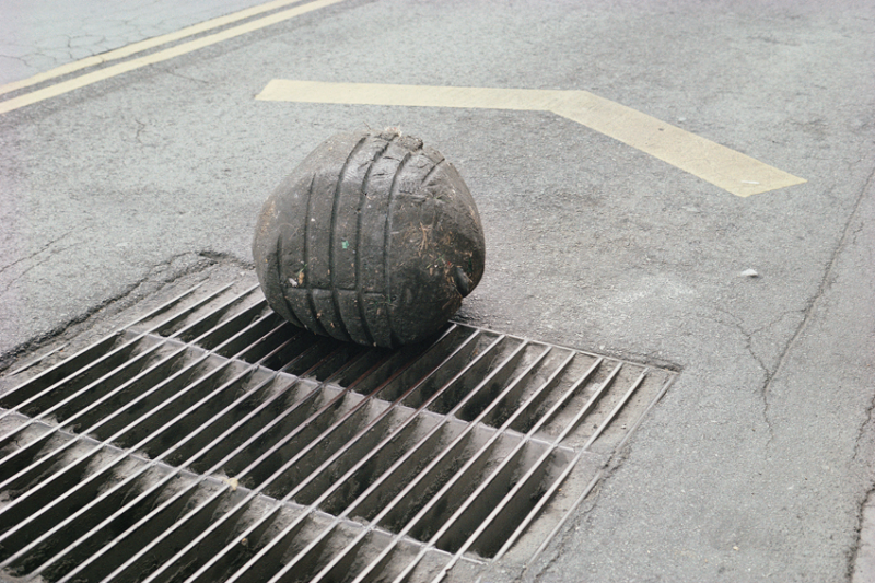 A color photograph of a large, grey plasticine sphere on a dirty grate in the middle of a paved street. The sphere shows indentations from the grate.