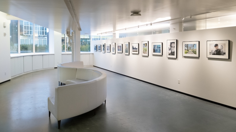 A gallery space with windows at one end, an s-shaped white sofa, and a dozen framed photographs hanging on a long white wall.