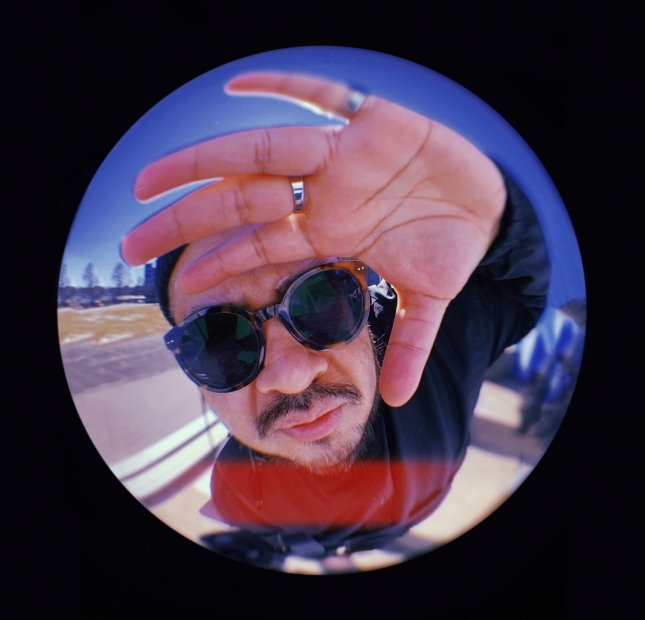 A fish-eye lens photo of a man wearing sunglasses with his hands up covering part of his face.