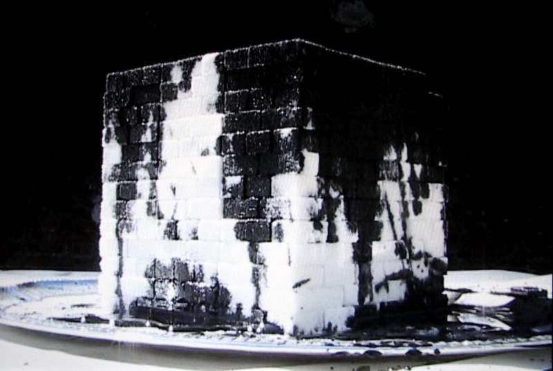 A video still of a stack of sugar cubes covered in dark oil on a silver plate.