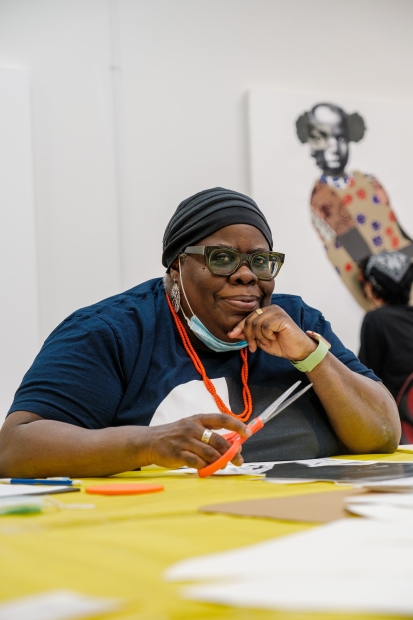 A Black woman pauses from cutting images out at a table