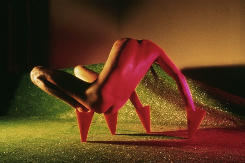 A color photograph of the artist, a light-skinned man, crouching and balancing, nude, on inverted orange cones on his hands and feet, in a field of glitter. 