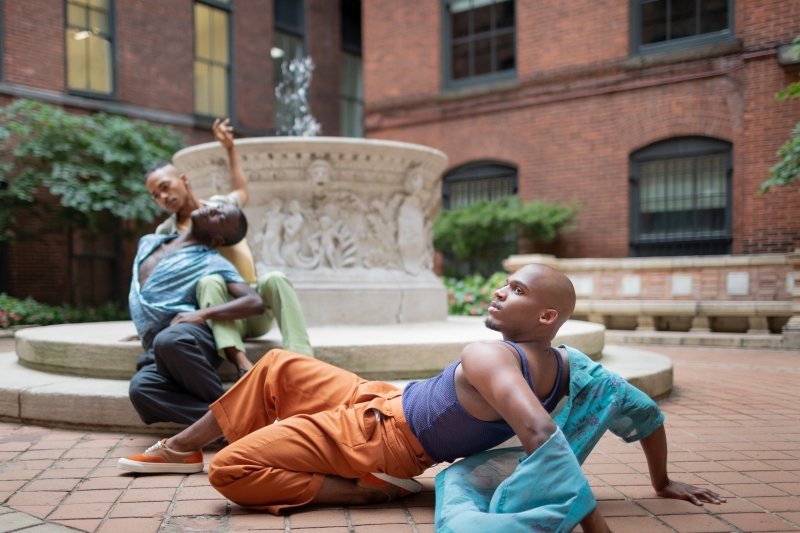 Three performers dressed in jewel tones posing sensually on the ground of a courtyard