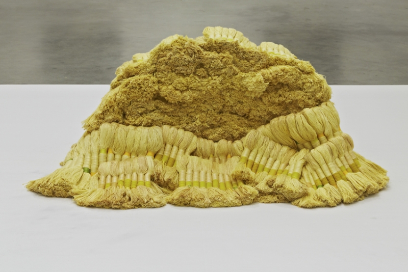 A sculpture made of bundles of yellow-colored linen and wool thread piled on a large white platform.