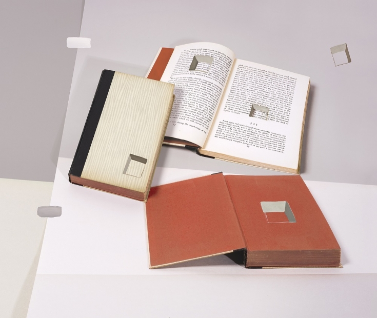 A color photograph depicts two open books and one closed book with illusionistic square holes painted on their surfaces and laid on top of creased paper supports to appear as though a still life in a flat or two-dimensional plane.