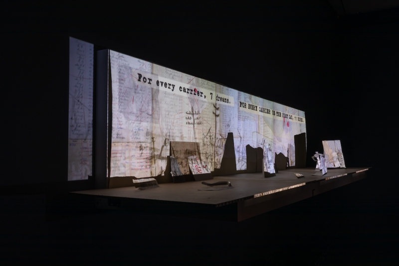 A close up of miniature paper set pieces, and a projection featuring a collage of letters and documents, drawings, and the words in typewriter "For every carrier, 7 dreams."