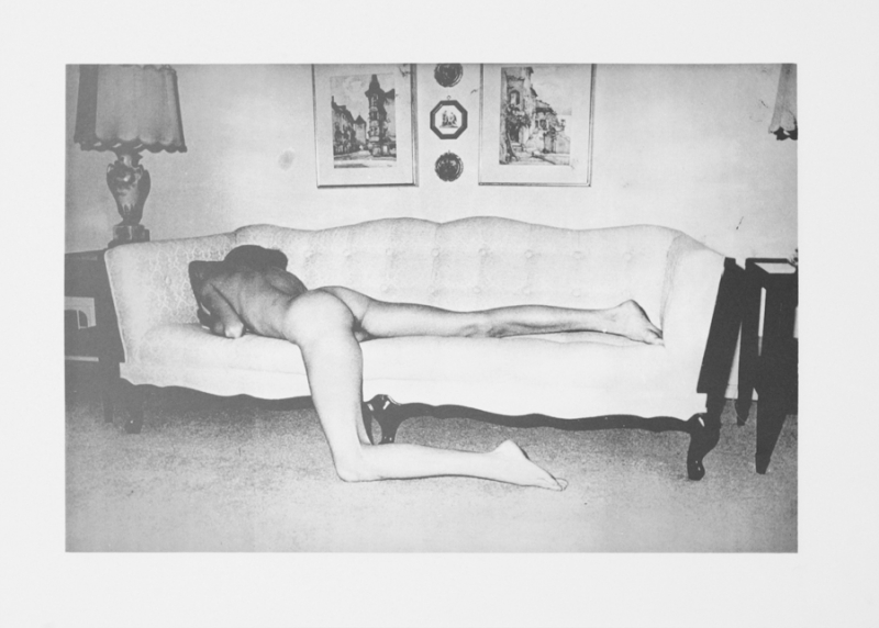 A grainy black-and-white photograph of a pale nude figure lounging face-down on a couch, their left leg hanging down and resting on the floor. 