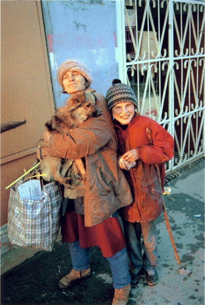 A color photograph shows a pale older woman holding a dog and a plastic bag of various materials, standing next to a pale school-age boy in a red jacket and black patterned knit hat who smiles directly at the viewer.