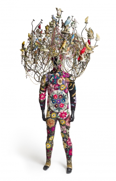 A sculpture composed of a colorful costume covering the body of a mannequin a chandelier-like headpiece decorated with ceramic birds and strings of beads.