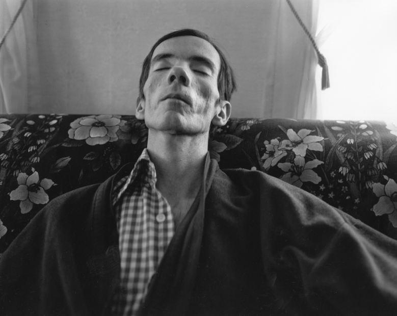 A black-and-white photograph of a pale, gaunt man in a bathrobe laying his head back as he sits on a floral couch.