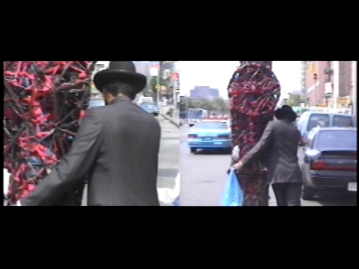 Two side-by-side video stills show artist Nari Ward from behind as he pushes a large, towering sculpture on wheels down a crowded street wearing a suit and hat.
