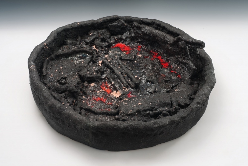 A matte black ceramic object in the shape of a shallow dish with unidentifiable contents and a scattering of red accents.