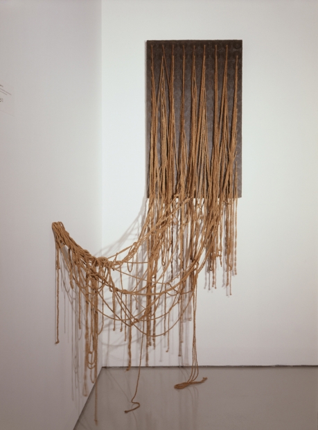 An installation of ochre- colored string attached in a regular pattern to a piece of grey painted plywood but tangling as they fall to the floor or are attached loosely to the adjacent wall.