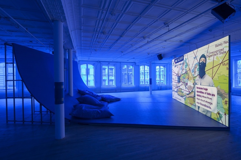 An installation photo cast in blue light of a large video screen showing a masked man in front of a map of Europe, opposite a curved wall-to-ceiling quarter pipe with lounge seating.