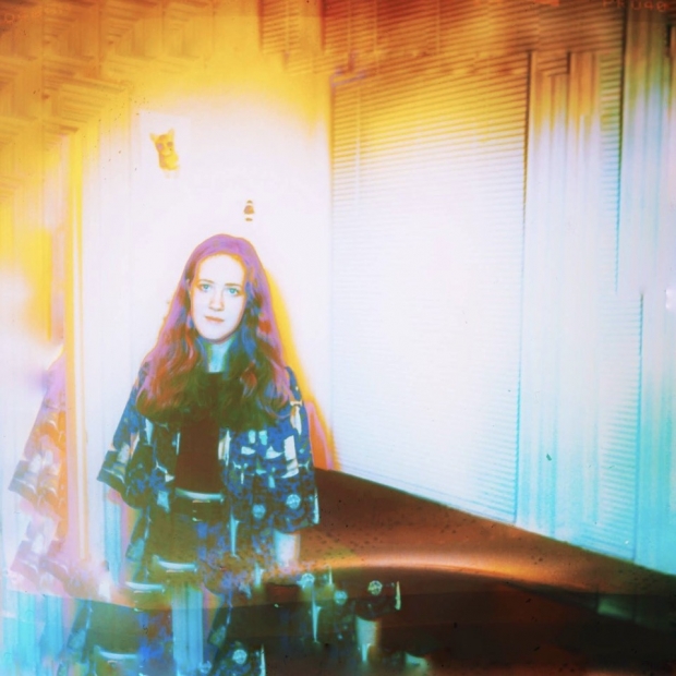 A color distorted, high-exposure photograph of a fair skin person with long way hair in a corner of a room.