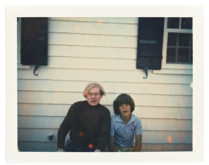 A Polaroid of Andy Warhol and preadolescent John F. Kennedy Jr. sitting close together in front of a white clapboard house.