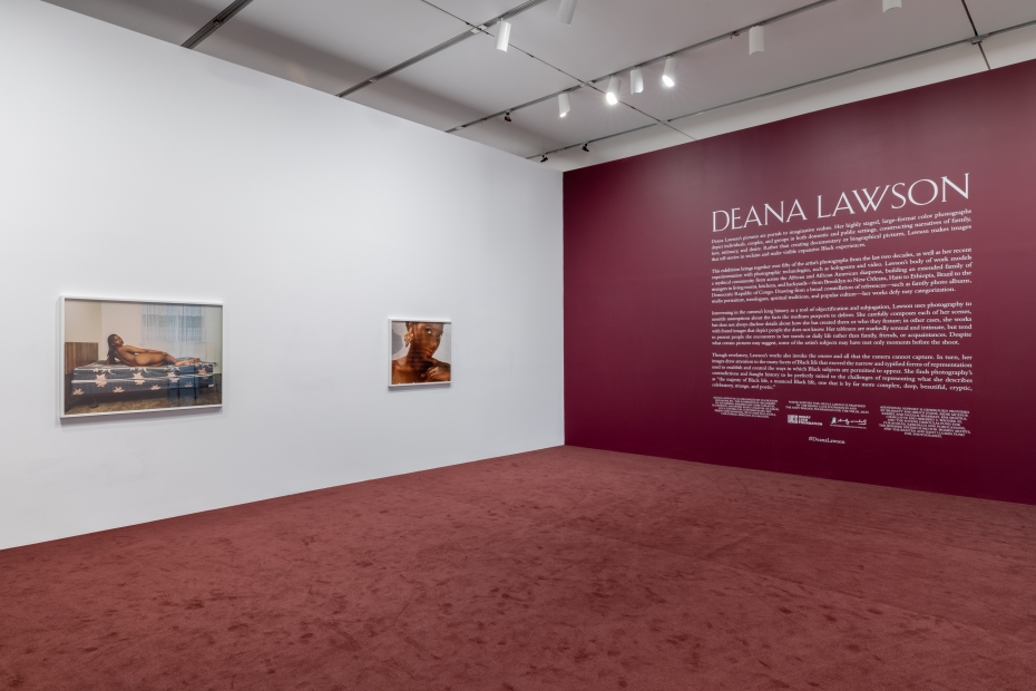 Two framed photos of Black women hang on a white wall opposite a painted burgundy wall with white text and "Deana Lawson" in big letters. The floor is carpeted in the same color.