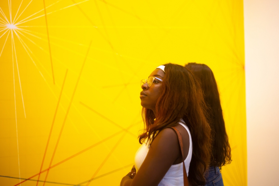 A young woman with dark hair and skin obscures a second in front of a bright yellow artwork.