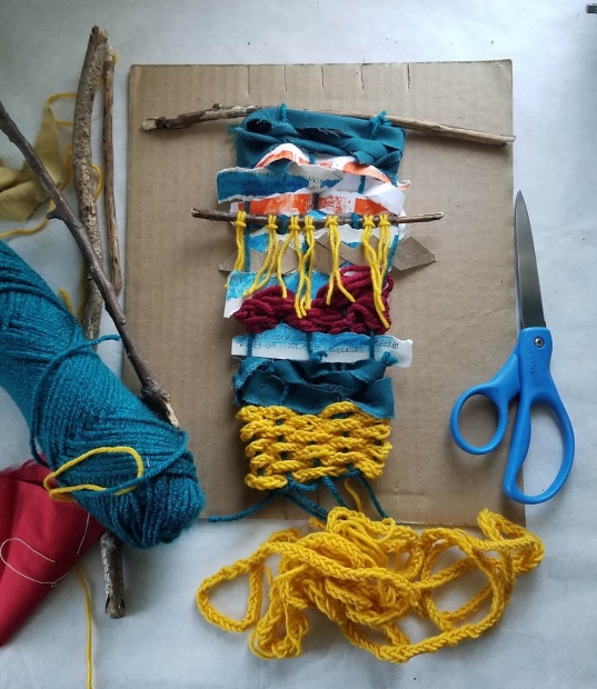 Scraps of blue yawn, fabric, paper, twigs woven together over cardboard with scissors on the side.