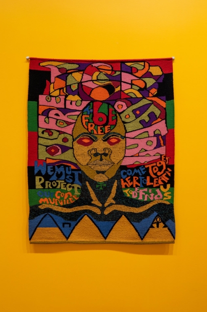 A colorful textile depicting a face with brown skin and red eyes surrounded by words including "To be free," hanging on an orange wall.