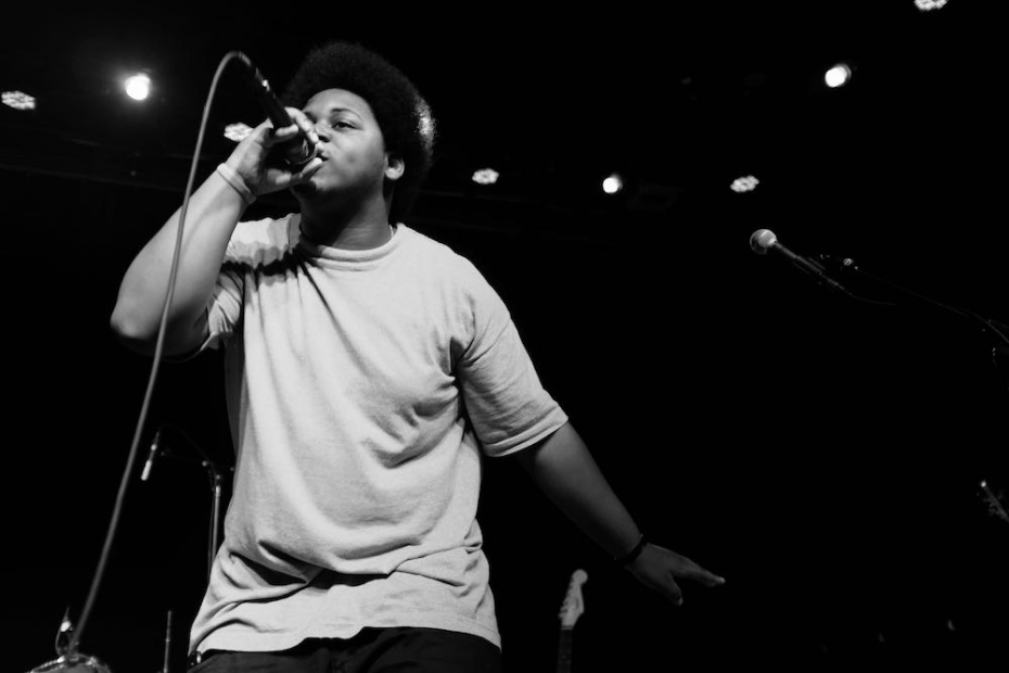 A black and white photo shows a young man performing on stage, holding a microphone, against a black background.