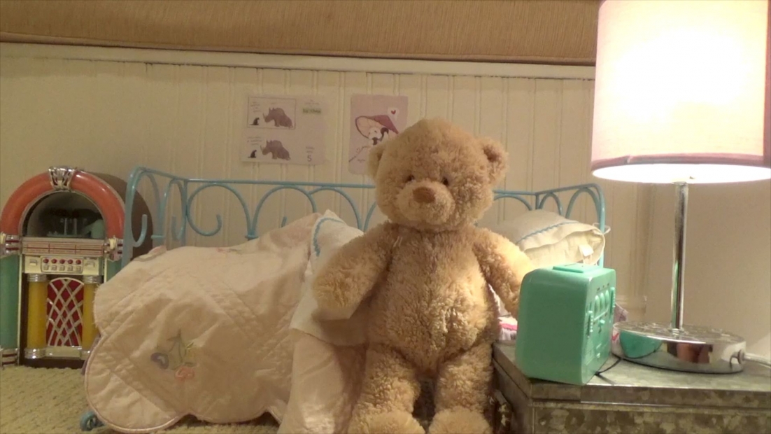 A stuffed bear propped up next to a night stand in a bedroom