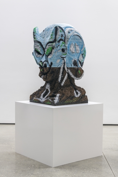 A bust by Huma Bhaba, that is gray and organic in texture. The top is light blue, and the figure does not appear to be human. 
