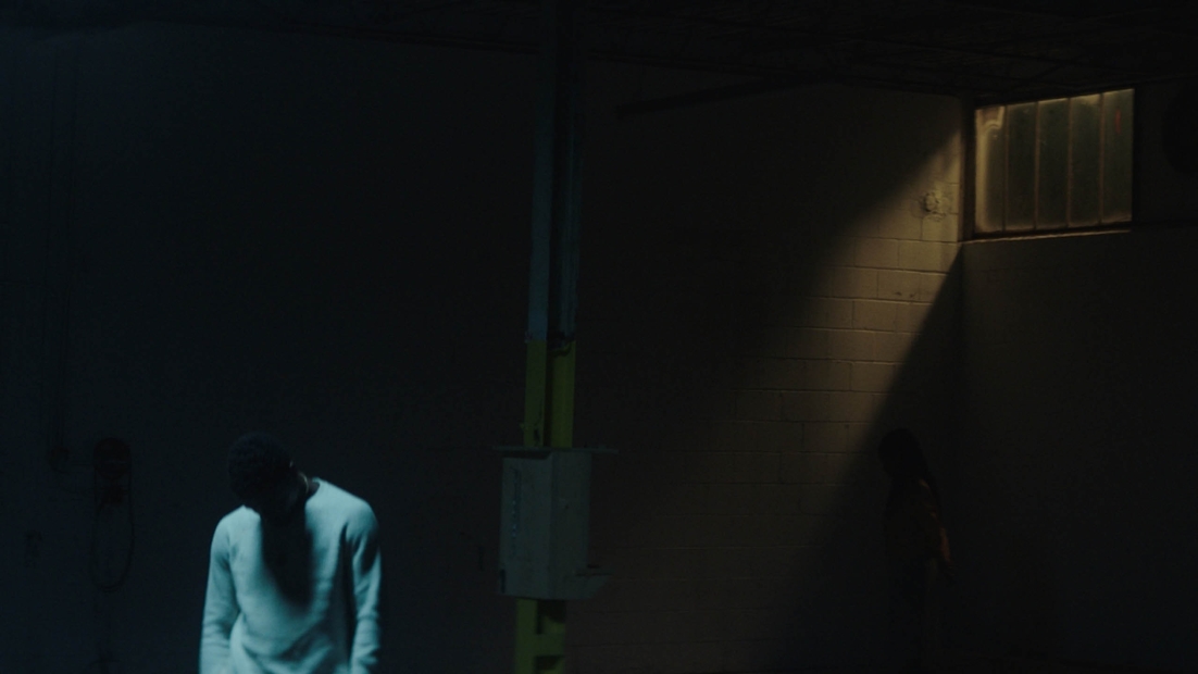 Dimly lit scene of a man in a white sweater standing in a dark room lit only by a window with bars.