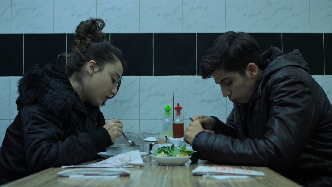 Two young people wearing coats eat at a restaurant with tiled walls