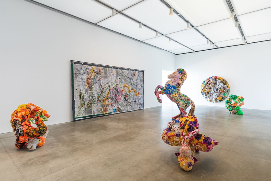 Installation shot showing large multi-colored bejeweled sculptures and a collaged tapestry on the wall. In the center of the room is a large bejeweled horse sculpture.